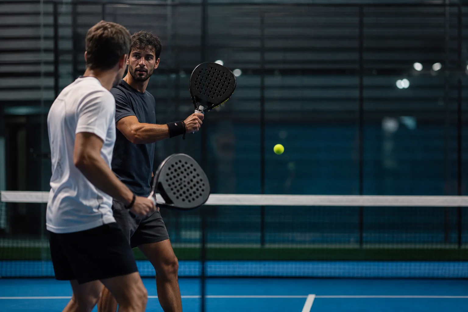 Two padel players at the net in a tense exchange, blurred glass wall behind them under court floodlights
